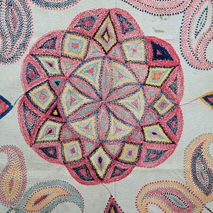Könnte beinhalten: Ein weißes Textil mit aufwendiger Handstickerei in einem zentralen kreisförmigen Mandalamuster aus rosa, gelben, blauen und dunkelblauen geometrischen Formen, umgeben von sich wiederholenden Paisley-Motiven und zarten floralen Rankenbordüren.