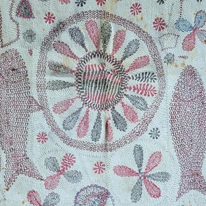 Op de afbeelding: Crèmekleurig textiel met ingewikkelde handgestikte borduursels. Voorzien van twee gestileerde vissen, een centraal cirkelvormig ontwerp en bloemen- en stermotieven in rood, zwart en blauw. De steken creëren een gedetailleerd patroon.