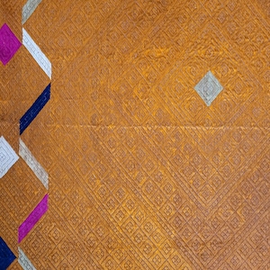 Op de afbeelding: Een mosterdgele stof met een herhalend rasterpatroon van kleine vierkantjes. Een verticale rand aan de linkerkant toont een zigzagontwerp in afwisselende kleuren donkerblauw, magenta, wit en lichtgrijs, met een bijpassende horizontale afwerking.