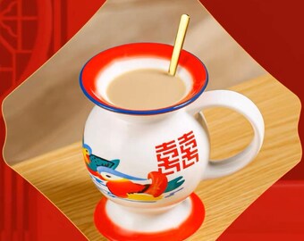 Puede incluir: Taza de cerámica blanca con un diseño rojo, azul y amarillo que presenta un pájaro y el carácter chino para "felicidad". La taza tiene un asa y una base redonda. Está llena de un líquido marrón claro y una cuchara dorada está dentro.