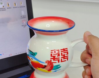 Puede incluir: Una taza de cerámica blanca con un diseño rojo, azul y amarillo. La taza tiene un asa y una base ancha. La taza tiene un carácter chino en la parte delantera que dice "宮".