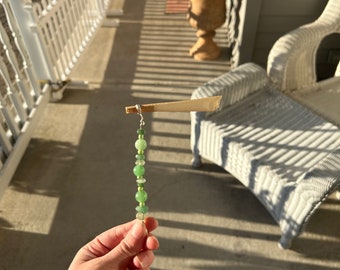 May include: A hand holds a green beaded necklace with a small, brown paper cone attached to the top. The necklace is made of round, green beads with a silver wire connecting them. The background is a blurry image of a white picket fence and a white wicker chair.