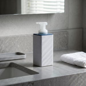 May include: A rectangular soap dispenser with a gray marble-like pattern. The dispenser has a blue top and a clear pump. A white towel and a small tray are also visible on the countertop.