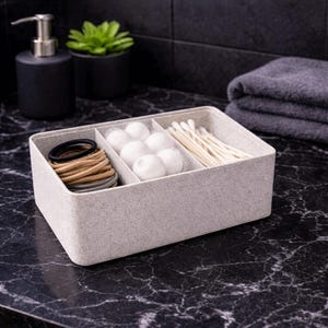 Puede incluir: Un organizador rectangular gris claro con cuatro compartimentos. Los compartimentos contienen gomas para el pelo, bolas de algodón y bastoncillos. El organizador está sobre una encimera de mármol negro, con un dispensador de jabón negro y una pequeña planta en maceta.