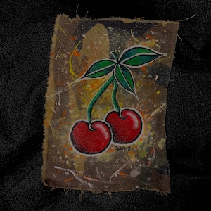 May include: A hand-painted artwork on a piece of fabric features two red cherries with green leaves and stems. The background has a splattered paint effect with shades of brown, yellow, and white. The artwork is set against a black background.