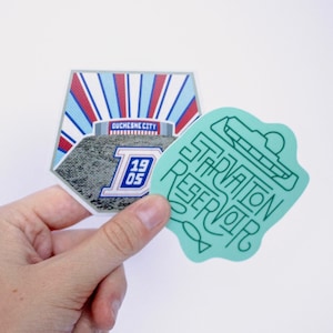 May include: Two stickers, one with a green outline of a reservoir and the text "Starvation Reservoir" and the other with a gray and blue illustration of a stadium with the text "Ouchesne City 1905".
