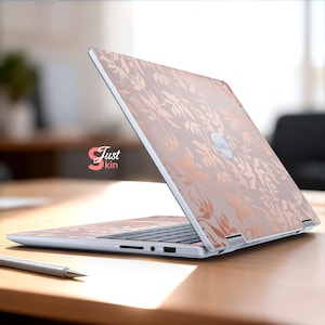 May include: A Dell laptop with a floral design skin in rose gold and light pink. The laptop is open on a wooden table, with a pen in the foreground. The "Just kin" logo is visible on the side of the laptop.