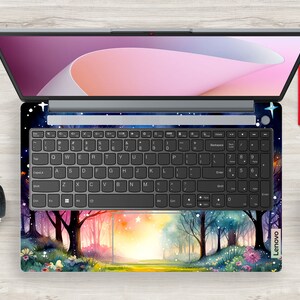 May include: A laptop computer with a colorful forest scene design on the keyboard and trackpad. The design features trees, flowers, and a path leading to a bright light. The laptop screen is closed and shows a pink and white abstract design.