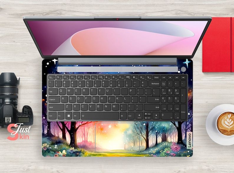 May include: A laptop computer with a colorful forest scene design on the keyboard and trackpad. The design features trees, flowers, and a path leading to a bright light. The laptop screen is closed and shows a pink and white abstract design.