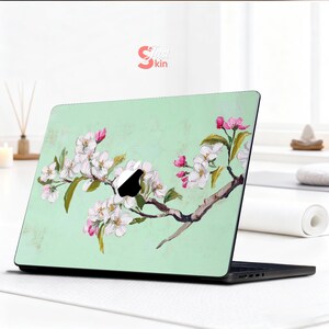May include: Laptop skin with a floral design. White and pink blossoms adorn a brown branch set against a light green backdrop. The skin has the text "Just Skin" in the upper right corner.