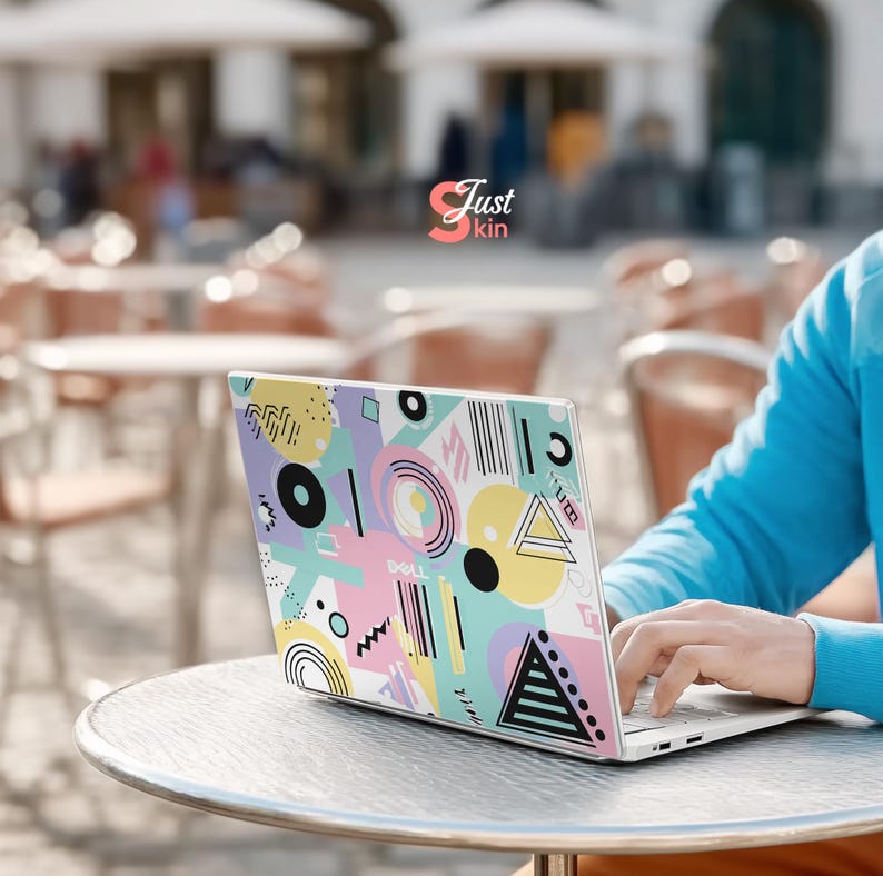 May include: A Dell laptop with a colorful geometric design. The design features shapes like circles, triangles, and lines in pastel colors including pink, yellow, and blue. The laptop is on a round table.