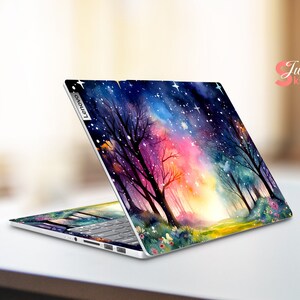May include: A laptop computer with a colorful watercolor design on the lid. The design features a forest scene with trees, a path, and a starry sky. The laptop is open and the keyboard is visible. The laptop brand is Lenovo.
