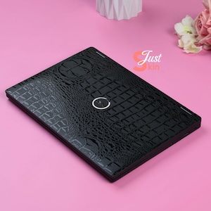 Pode incluir: Capa de laptop preta com textura de crocodilo. A capa tem um recorte circular para o logotipo do laptop e o texto "Just kin" no canto. A capa do laptop está em uma superfície rosa.