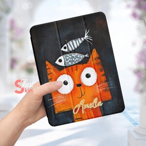 May include: A black and orange tablet case featuring a cartoon illustration of a cat with large eyes and two fish on top of its head. The cat's name, "Amelia", is written in gold lettering on the case.