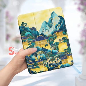 Könnte beinhalten: Eine gelbe und blaue iPad-Hülle mit japanisch inspiriertem Design, das Berge, Bäume und Gebäude zeigt. Der Name "Amelia" ist in goldener Schrift auf der Hülle geschrieben.