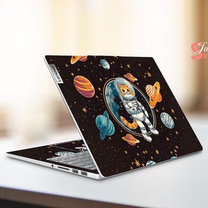 May include: A Lenovo laptop with a black skin featuring a cartoon cat astronaut floating in space. The design includes planets, stars, and a blue and orange colour scheme. The laptop is open, displaying the keyboard.