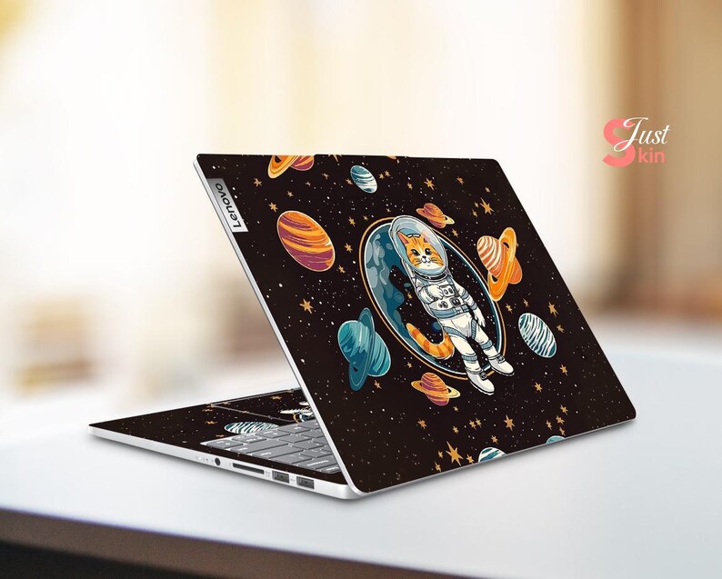 May include: A Lenovo laptop with a black skin featuring a cartoon cat astronaut floating in space. The design includes planets, stars, and a blue and orange colour scheme. The laptop is open, displaying the keyboard.