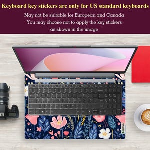 Op de afbeelding: Laptop met een bloemen toetsenbord skin met roze en witte bloemen, blauwe bladeren en een marineblauwe achtergrond. Het laptopscherm toont een roze en witte gradi&euml;nt. Een camera en een kopje koffie zijn zichtbaar.