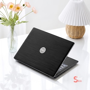 Peut inclure: Un ordinateur portable Dell noir avec une texture en métal brossé sur le couvercle, arborant un logo Dell circulaire argenté au centre. L'ordinateur est entrouvert, révélant une partie du clavier, et est posé sur une surface blanche.