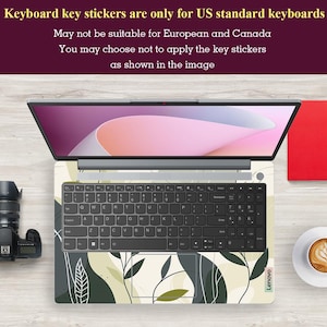 May include: A laptop computer with a keyboard covered in green and white floral key stickers. The laptop is on a wooden surface with a camera and a cup of coffee.