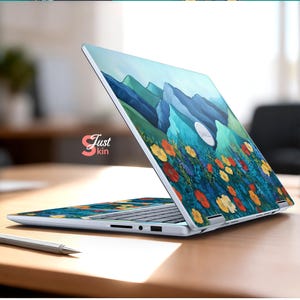 Pode incluir: Um laptop com um design floral e de montanha. O design apresenta flores coloridas em primeiro plano e montanhas azuis ao fundo. O laptop est&aacute; aberto sobre uma superf&iacute;cie de madeira. O texto "Just kin" &eacute; vis&iacute;vel.