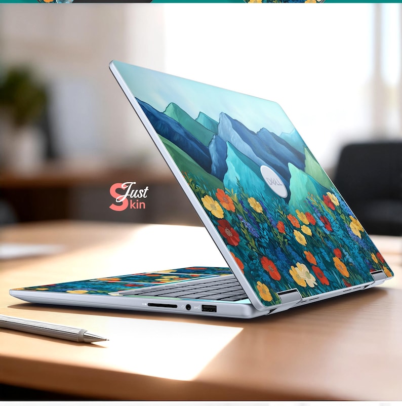Pode incluir: Um laptop com um design floral e de montanha. O design apresenta flores coloridas em primeiro plano e montanhas azuis ao fundo. O laptop est&aacute; aberto sobre uma superf&iacute;cie de madeira. O texto "Just kin" &eacute; vis&iacute;vel.