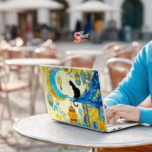 May include: Laptop with a decorative skin featuring a black cat silhouette perched on a rooftop with a crescent moon and lanterns. The design incorporates yellow and blue watercolor elements. The text "Just kin" is visible.