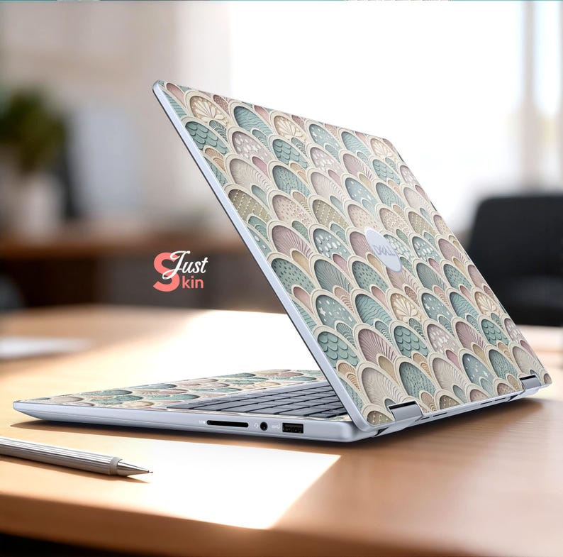 Puede incluir: Un port&aacute;til con una funda decorativa que presenta un patr&oacute;n repetido de formas estilizadas y superpuestas en tonos beige, azul y rosa. El port&aacute;til est&aacute; abierto sobre una superficie de madera. El texto "Just kin" es visible.