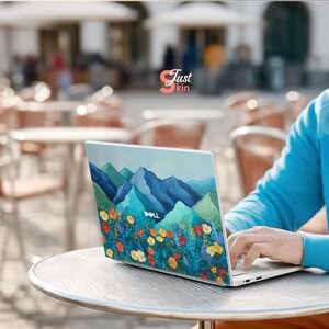 Pode incluir: Um laptop Dell com um design de montanha e floral na tampa. O laptop est&aacute; aberto em uma mesa, com uma pessoa digitando no teclado. O design apresenta montanhas azuis e flores coloridas.