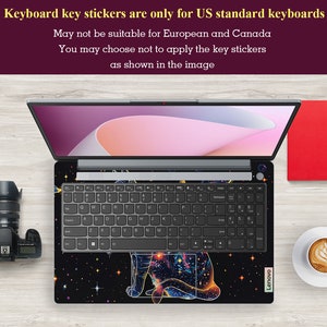 May include: A black laptop with a galaxy-themed keyboard sticker featuring a cat silhouette. The sticker has a blue, yellow, and orange color scheme with a starry background. The text "Lenovo" is visible on the bottom right corner of the laptop.