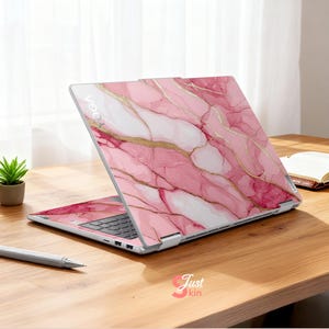 Op de afbeelding: Een laptop met een roze marmerpatroon en gouden accenten. De laptop is open en toont het toetsenbord en het scherm. Het woord "YOGA" is zichtbaar aan de bovenkant. Het staat op een houten bureau met een pen en een kleine plant.