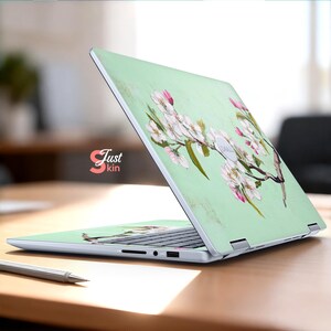 May include: A laptop skin with a floral design of white and pink blossoms on a pale green background. The laptop is open on a wooden desk. The text "Just kin" is visible.