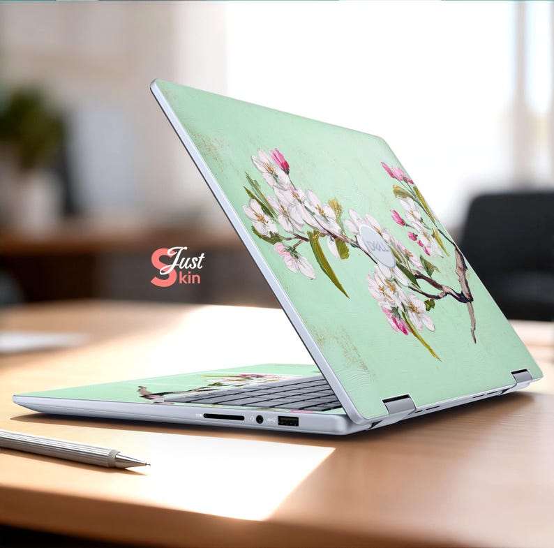 May include: A laptop skin with a floral design of white and pink blossoms on a pale green background. The laptop is open on a wooden desk. The text "Just kin" is visible.