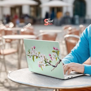 May include: A laptop with a light green cover featuring a floral design of white blossoms and pink buds on a brown branch. The laptop is on a round table, and the text "Just kin" is visible.