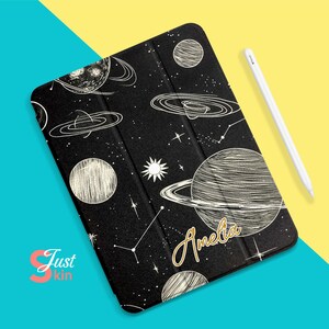 Puede incluir: Funda negra para tableta con diseño de galaxia blanca, que incluye planetas, estrellas y constelaciones. El nombre "Amelia" está escrito en oro. Un lápiz óptico blanco está al lado de la funda. Fondo azul y amarillo.
