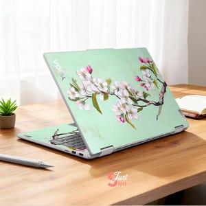 May include: A laptop with a light green cover featuring a floral design of white and pink blossoms on a brown branch. The laptop is open on a wooden table with a pen and small plant.