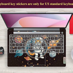 May include: Laptop with a keyboard skin featuring an astronaut in space, planets, and stars. The keyboard keys have colourful illustrations. The text "Keyboard key stickers are only for US standard keyboards" is visible. The laptop is on a wooden surface.
