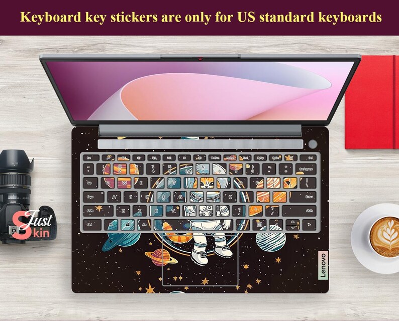 May include: Laptop with a keyboard skin featuring an astronaut in space, planets, and stars. The keyboard keys have colourful illustrations. The text "Keyboard key stickers are only for US standard keyboards" is visible. The laptop is on a wooden surface.