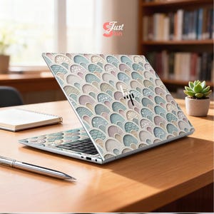 Puede incluir: Un ordenador port&aacute;til con una cubierta decorativa que presenta un patr&oacute;n repetido de formas estilizadas y superpuestas en colores pastel. El port&aacute;til est&aacute; abierto sobre un escritorio de madera, con un bol&iacute;grafo y un cuaderno cerca. El logotipo de HP es visible.