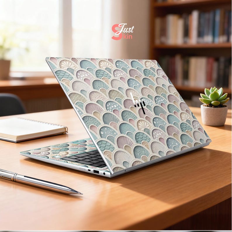 Puede incluir: Un ordenador port&aacute;til con una cubierta decorativa que presenta un patr&oacute;n repetido de formas estilizadas y superpuestas en colores pastel. El port&aacute;til est&aacute; abierto sobre un escritorio de madera, con un bol&iacute;grafo y un cuaderno cerca. El logotipo de HP es visible.