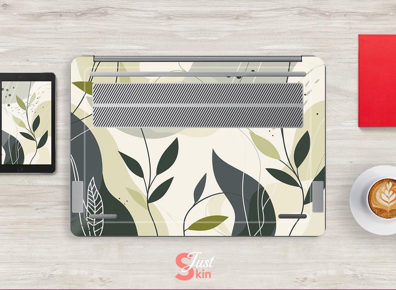 May include: Laptop skin with a white background and a pattern of green and gray leaves. The skin has a silver metal vent in the center.