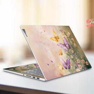 Puede incluir: Un ordenador port&aacute;til con un dise&ntilde;o floral rosa, blanco y amarillo con mariposas. El port&aacute;til tiene un logotipo de Lenovo plateado en la parte posterior.