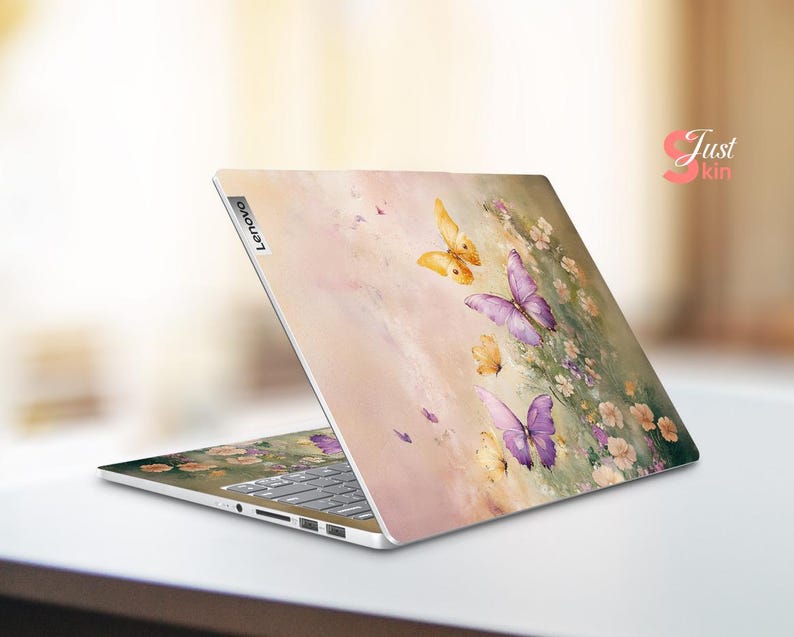 Puede incluir: Un ordenador port&aacute;til con un dise&ntilde;o floral rosa, blanco y amarillo con mariposas. El port&aacute;til tiene un logotipo de Lenovo plateado en la parte posterior.