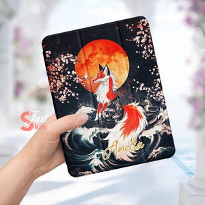 May include: A black phone case featuring a red moon, white cherry blossom branches, and a red fox with a long tail. The fox is surrounded by blue waves and the name "Amelia" is written in gold lettering.