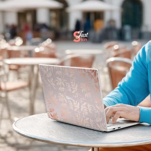 May include: A silver laptop with a floral design in rose gold is open on a round table. The laptop is being used outdoors. The text "Just kin" is in the upper left corner of the image.
