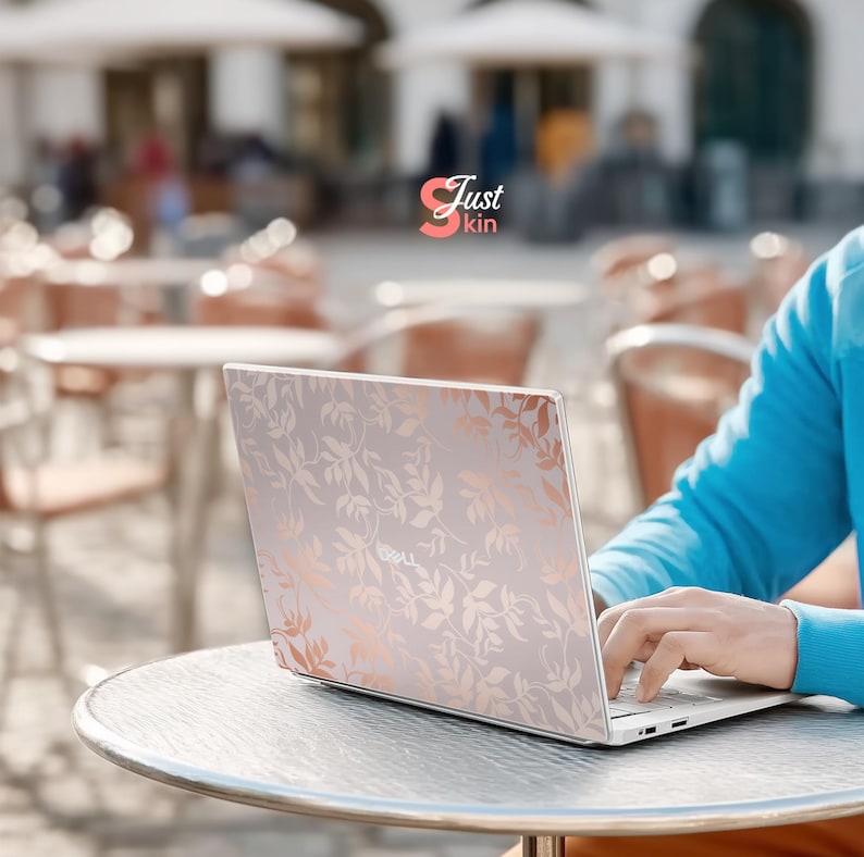 May include: A silver laptop with a floral design in rose gold is open on a round table. The laptop is being used outdoors. The text "Just kin" is in the upper left corner of the image.