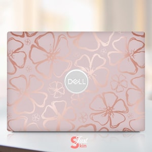 Pode incluir: Uma capa para laptop Dell com um padrão floral em ouro rosa sobre um fundo rosa claro. O design inclui contornos de flores grandes e pequenas. O logotipo da Dell está centralizado. As palavras "Just skin" estão na parte inferior.