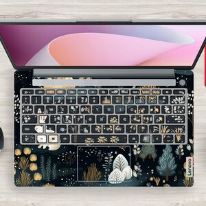 May include: A laptop with a forest-themed keyboard skin, featuring trees, plants, and a bear illustration. The keyboard keys are black with white and gold designs. The laptop screen is open, showing a pink and purple display.