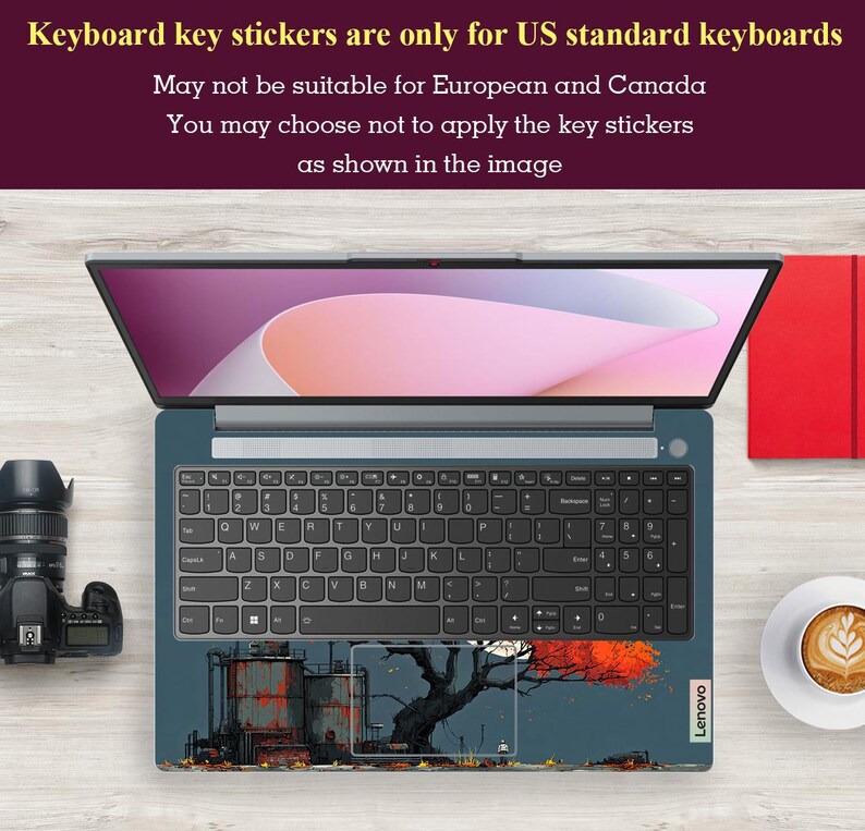 May include: An open Lenovo laptop with a keyboard skin featuring an industrial landscape design. The screen displays a pink and white gradient. A camera and a cup of coffee are visible. Text at the top states: "Keyboard key stickers are only for US standard keyboards."