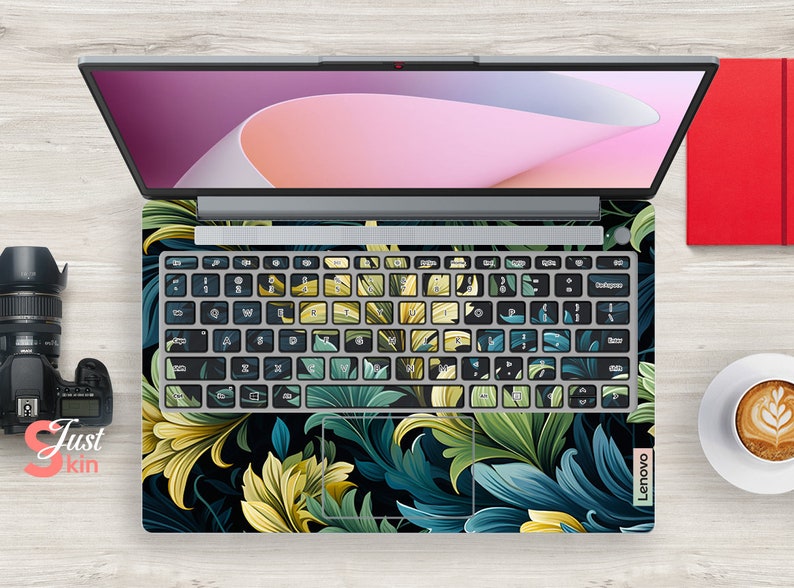 May include: A laptop computer with a floral design keyboard skin. The skin features a dark background with green and yellow leaves.
